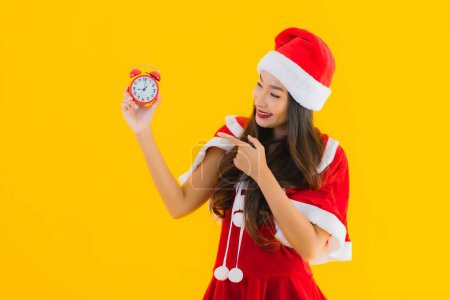 Téléchargez les photos : Portrait belle jeune femme asiatique porter des vêtements de Noël et chapeau montre horloge ou alarme sur fond isolé jaune - en image libre de droit