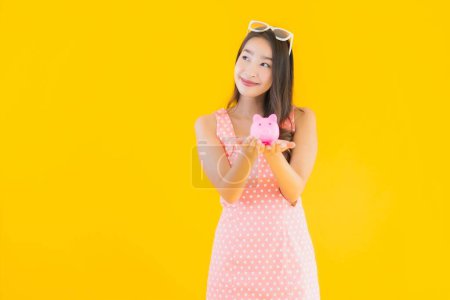 Téléchargez les photos : Portrait belle jeune femme asiatique montrer beaucoup d'argent ou d'argent avec tirelire sur fond jaune isolé - en image libre de droit
