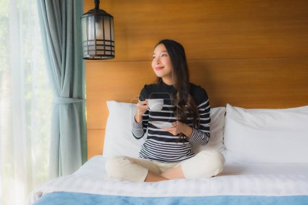 Foto de Retrato joven mujer asiática en la cama en el interior del dormitorio con taza de café blanco - Imagen libre de derechos