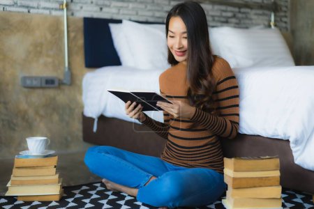 Foto de Joven mujer asiática con taza de café y leer libro en la cama en el interior del dormitorio - Imagen libre de derechos