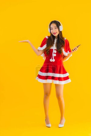 Téléchargez les photos : Portrait belle jeune asiatique vêtements de Noël et chapeau écouter de la musique avec casque et smartphone sur fond isolé jaune - en image libre de droit