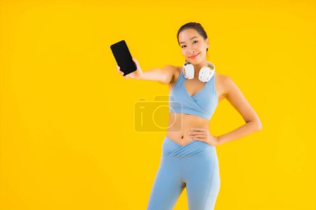 Téléchargez les photos : Portrait belle jeune femme asiatique porter des vêtements de sport avec téléphone intelligent et casque pour écouter de la musique sur fond jaune - en image libre de droit