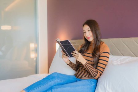 Téléchargez les photos : Portrait belle jeune femme asiatique lire livre sur lit dans chambre à coucher intérieur - en image libre de droit