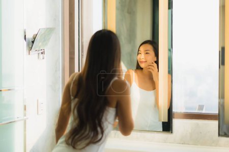 Téléchargez les photos : Portrait belle jeune femme asiatique vérifier son visage et le visage dans la salle de bain intérieure - en image libre de droit