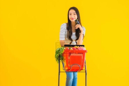 Téléchargez les photos : Portrait belle jeune asiatique femme shopping épicerie chariot de supermarché avec carte de crédit sur fond jaune isolé - en image libre de droit