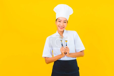 Photo for Portrait beautiful young asian woman wear chef or cook uniform with spoon and fork on yellow isolated background - Royalty Free Image