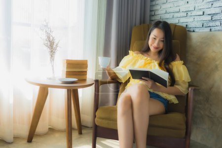 Téléchargez les photos : Portrait jeune asiatique femme assis sur canapé chaise et lire livre avec tasse de café dans salon intérieur - en image libre de droit