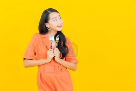 Téléchargez les photos : Portrait belle jeune femme asiatique sourire avec cuillère et fourchette sur fond de couleur jaune - en image libre de droit