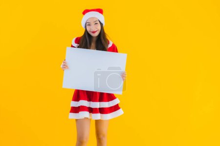 Téléchargez les photos : Portrait belle jeune femme asiatique vêtements de Noël et chapeau sourire avec panneau blanc vide sur fond isolé jaune - en image libre de droit