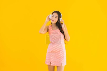 Foto de Retrato hermosa joven asiática mujer escuchar música con el teléfono móvil inteligente y auriculares en amarillo aislado fondo - Imagen libre de derechos