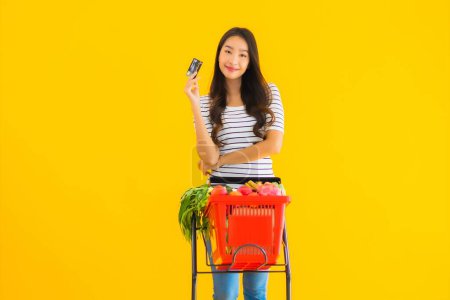 Foto de Retrato hermosa joven asiática mujer compras carrito de comestibles de supermercado con tarjeta de crédito en amarillo aislado fondo - Imagen libre de derechos