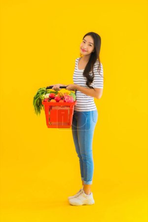 Téléchargez les photos : Portrait belle jeune femme asiatique avec panier d'épicerie panier du supermarché dans le centre commercial sur fond jaune isolé - en image libre de droit