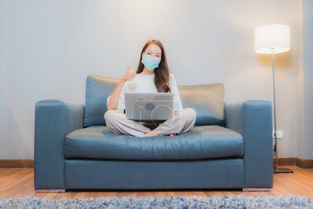 Téléchargez les photos : Portrait belle jeune femme asiatique porter un masque et utiliser ordinateur portable sur canapé dans le salon intérieur - en image libre de droit