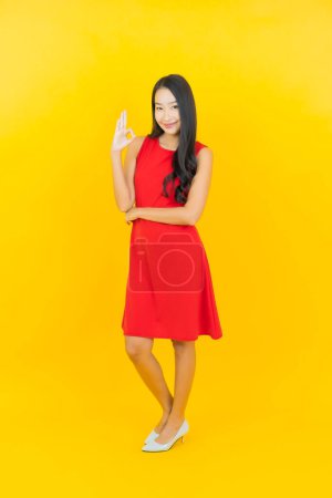Téléchargez les photos : Portrait belle jeune femme asiatique porter robe rouge sourire avec action sur fond de couleur jaune - en image libre de droit