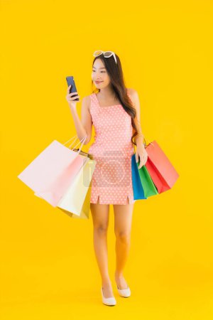Téléchargez les photos : Portrait belle jeune femme asiatique avec sac à provisions coloré avec téléphone portable intelligent et carte de crédit sur fond jaune isolé - en image libre de droit