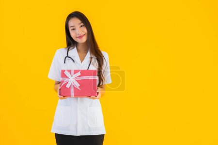 Téléchargez les photos : Portrait belle jeune asiatique médecin femme montrer boîte cadeau rouge sur fond jaune isolé - en image libre de droit