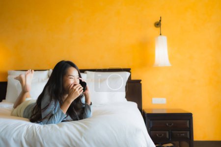 Photo for Portrait beautiful young asian woman using mobile cell phone on bed with coffee cup in bedroom interior - Royalty Free Image