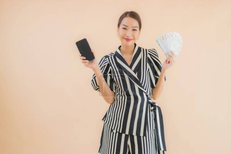 Foto de Retrato hermosa joven mujer asiática con dinero en efectivo y teléfono móvil inteligente sobre fondo marrón - Imagen libre de derechos