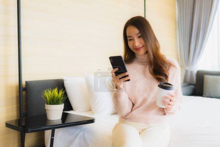 Photo for Portrait beautiful young asian woman with coffee cup and mobile phone on bed in bedroom interior - Royalty Free Image
