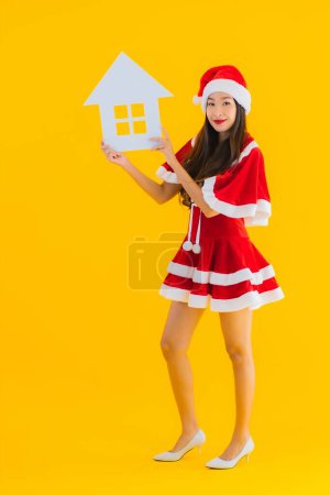 Photo for Portrait beautiful young asian woman wear christmas clothes and hat show home house sign with a lot of cash and money on yellow isolated background - Royalty Free Image