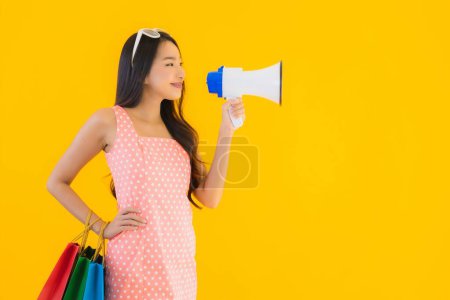 Téléchargez les photos : Portrait belle jeune femme asiatique avec mégaphone et sac à provisions du grand magasin sur fond jaune isolé - en image libre de droit