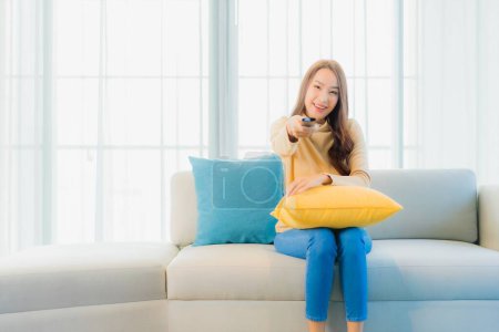 Foto de Retrato hermosa joven asiática mujer con control remoto tv en sofá en sala de estar interior - Imagen libre de derechos