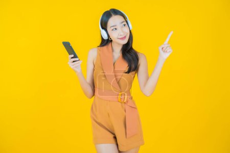 Téléchargez les photos : Portrait belle jeune femme asiatique avec casque et téléphone intelligent pour écouter de la musique sur fond jaune - en image libre de droit
