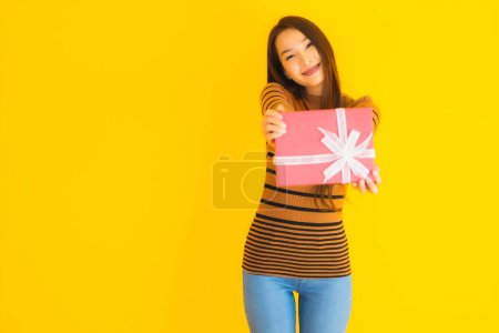 Téléchargez les photos : Portrait belle jeune femme asiatique sourire heureux avec boîte cadeau rouge sur fond jaune isolé - en image libre de droit