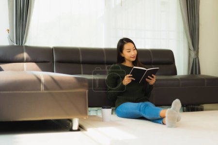 Téléchargez les photos : Portrait belle jeune femme asiatique lire livre avec tasse de café et canapé dans le salon intérieur - en image libre de droit