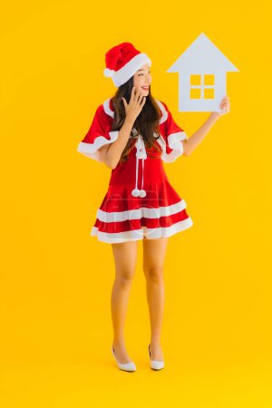 Photo for Portrait beautiful young asian woman wear christmas clothes and hat show home house sign with a lot of cash and money on yellow isolated background - Royalty Free Image