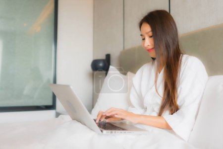 Téléchargez les photos : Portrait belle jeune femme asiatique utiliser un ordinateur portable ou un ordinateur dans la chambre à coucher intérieur - en image libre de droit