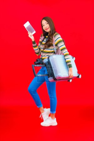 Téléchargez les photos : Portrait belle jeune femme asiatique avec bagages et carte d'embarquement sur fond rouge - en image libre de droit