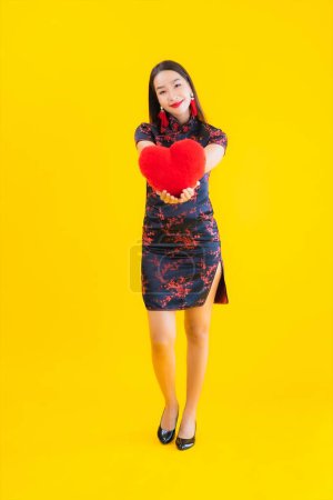 Téléchargez les photos : Portrait belle jeune femme asiatique porter robe chinoise montrer oreiller en forme de coeur sur fond jaune isolé - en image libre de droit
