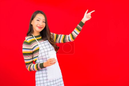 Foto de Retrato hermosa joven mujer asiática usar delantal con taza de café sobre fondo rojo - Imagen libre de derechos