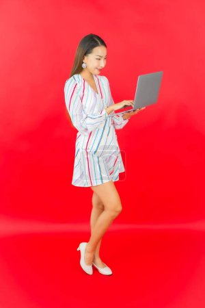 Téléchargez les photos : Portrait belle jeune femme asiatique utiliser ordinateur portable pour le travail sur fond rouge - en image libre de droit