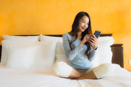Téléchargez les photos : Portrait belle jeune femme asiatique en utilisant un téléphone portable sur le lit avec tasse de café à l'intérieur de la chambre - en image libre de droit