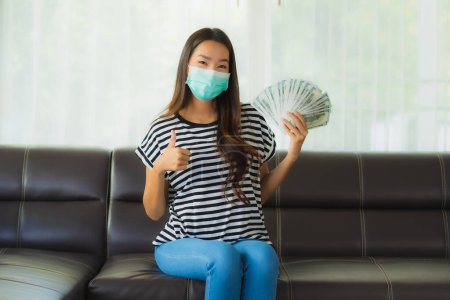 Photo for Portrait beautiful young asian woman with mask on sofa show money or cash at home - Royalty Free Image