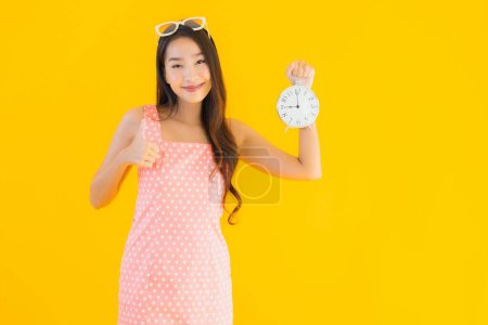 Téléchargez les photos : Portrait belle jeune femme asiatique montrer alarme ou horloge sur fond jaune isolé - en image libre de droit