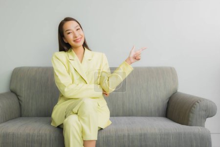 Téléchargez les photos : Portrait belle jeune femme asiatique assis avec le sourire sur le canapé dans le salon intérieur - en image libre de droit