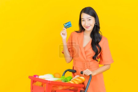 Téléchargez les photos : Portrait belle jeune femme asiatique sourire avec panier d'épicerie du supermarché sur fond de couleur jaune - en image libre de droit