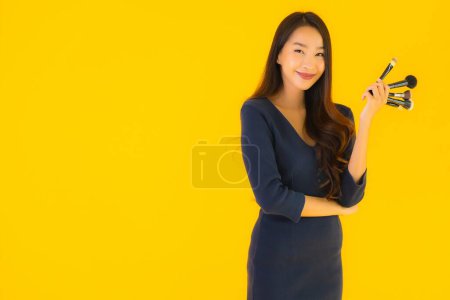 Téléchargez les photos : Portrait belle jeune femme asiatique avec pinceau de maquillage sur fond jaune isolé - en image libre de droit