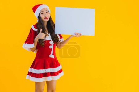 Foto de Retrato hermosa joven asiática mujer navidad ropa y sombrero sonrisa con vacío blanco tablero cartel sobre amarillo aislado fondo - Imagen libre de derechos