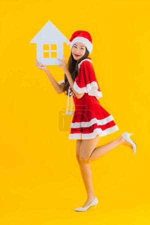 Foto de Retrato hermosa mujer asiática joven usar ropa de Navidad y sombrero mostrar casa casa signo con un montón de dinero en efectivo y dinero en amarillo aislado fondo - Imagen libre de derechos