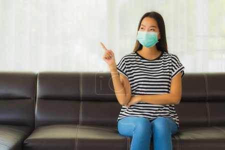 Téléchargez les photos : Portrait belle jeune femme asiatique avec masque rester à la maison sur canapé dans le salon pour protéger coronavirus ou covid19 - en image libre de droit