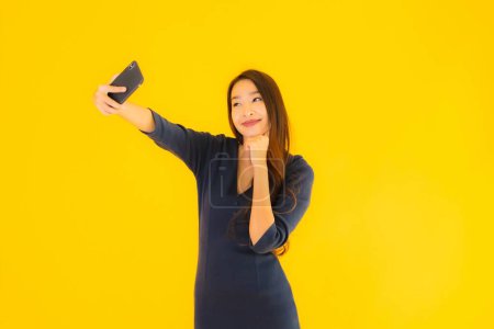 Téléchargez les photos : Portrait belle jeune femme asiatique avec téléphone mobile intelligent ou téléphone portable sur fond jaune isolé - en image libre de droit