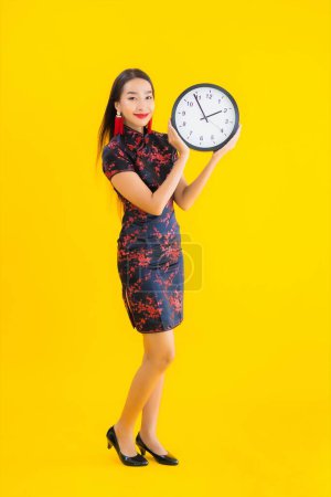 Photo for Portrait beautiful young asian woman wear chinese dress show clock or alarm on yellow isolated background - Royalty Free Image