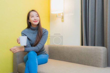 Téléchargez les photos : Portrait belle jeune femme asiatique sourire se détendre avec tasse de café sur canapé dans le salon intérieur - en image libre de droit