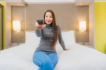 Téléchargez les photos : Portrait belle jeune femme asiatique utiliser la télévision à distance sur le lit pour changer de canal dans l'intérieur de la chambre - en image libre de droit