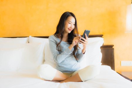 Foto de Retrato hermosa joven mujer asiática utilizando el teléfono celular móvil en la cama con taza de café en el interior del dormitorio - Imagen libre de derechos