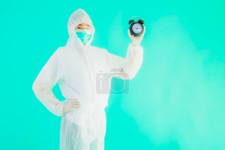 Photo for Portrait beautiful young asian doctor woman wear ppe suite and mask show clock or alarm on blue isolated background - Royalty Free Image